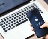 Hand hält Smartphone mit Schloss-Symbol und Passwort-Feld. Ein Laptop im Hintergrund erinnert an ein mögliches **Datenleck** und die Wichtigkeit eines sicheren **Passworts**.