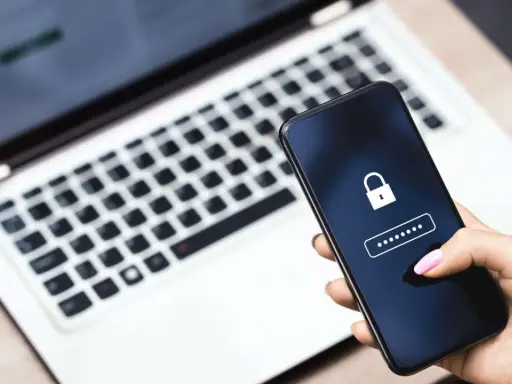 Hand hält Smartphone mit Schloss-Symbol und Passwort-Feld. Ein Laptop im Hintergrund erinnert an ein mögliches **Datenleck** und die Wichtigkeit eines sicheren **Passworts**.